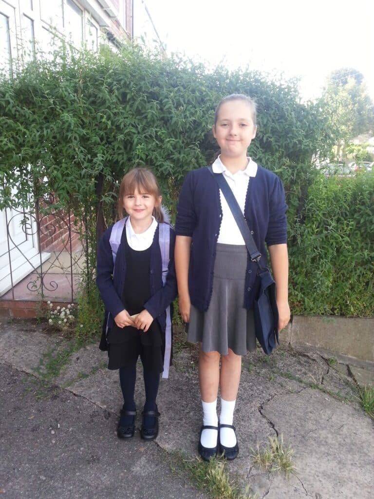 Two young girls stand outdoors on a path in front of green bushes. Both wear navy cardigans, white shirts, and skirts. The older girl is taller, wearing a grey skirt and white socks, smiling slightly. The younger girl wears a black pinafore and dark tights, smiling brightly.