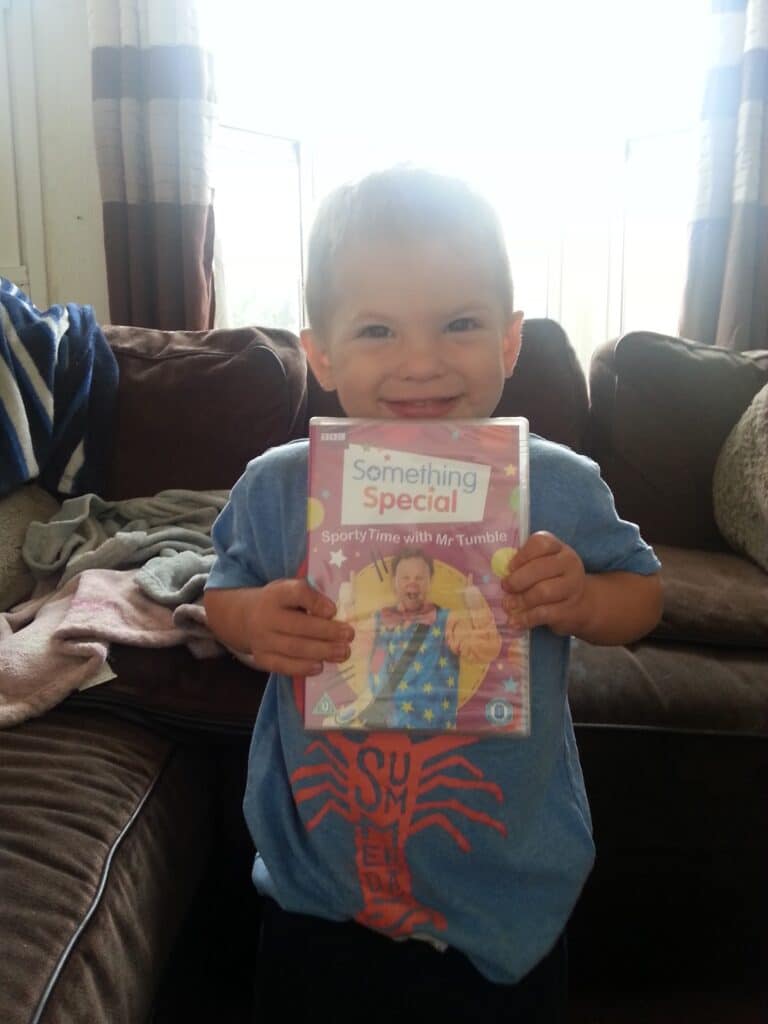 A smiling young child stands indoors, holding a DVD case of "Something Special: Sporty Time with Mr Tumble." The child wears a blue shirt and is in front of a brown sofa with blankets and cushions. Sunlight streams through large windows behind them.