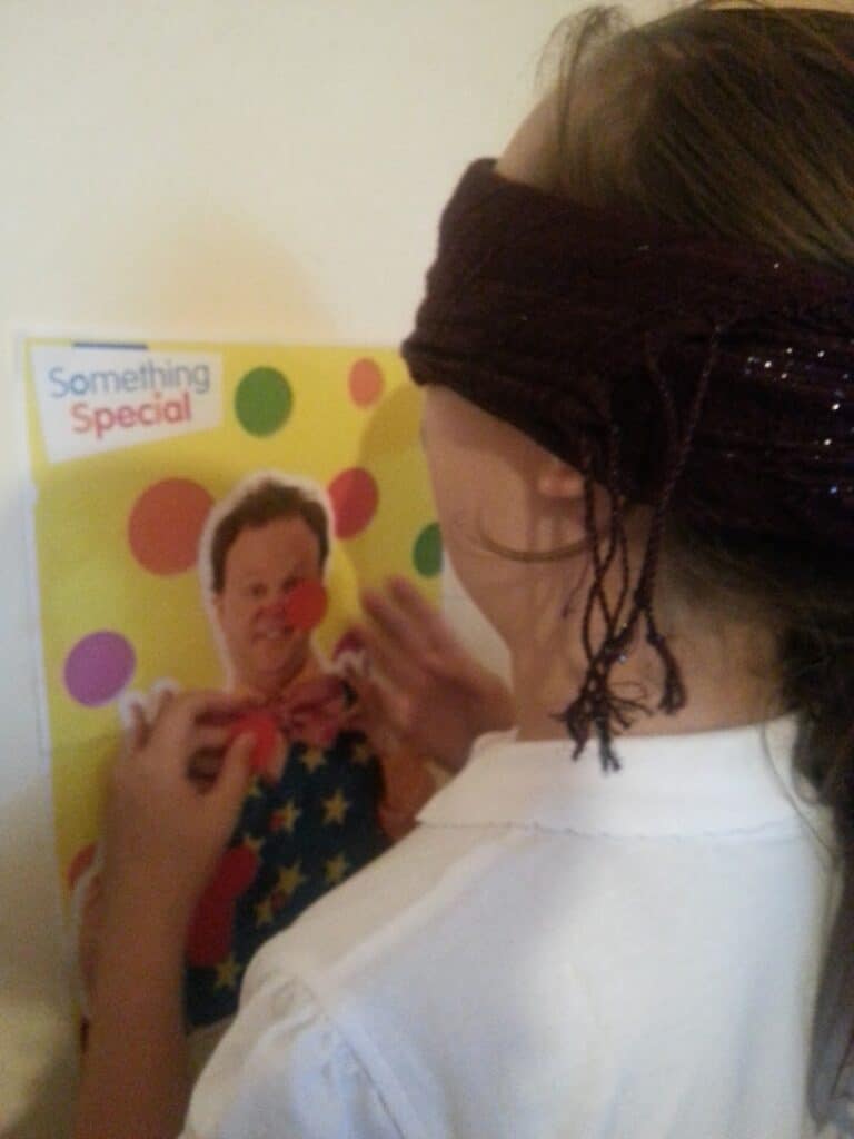 A child wearing a dark blindfold plays “pin the nose on the clown” using a colourful poster labelled "Something Special." The child is attempting to stick a red nose onto the clown’s face on the yellow, polka-dotted poster.