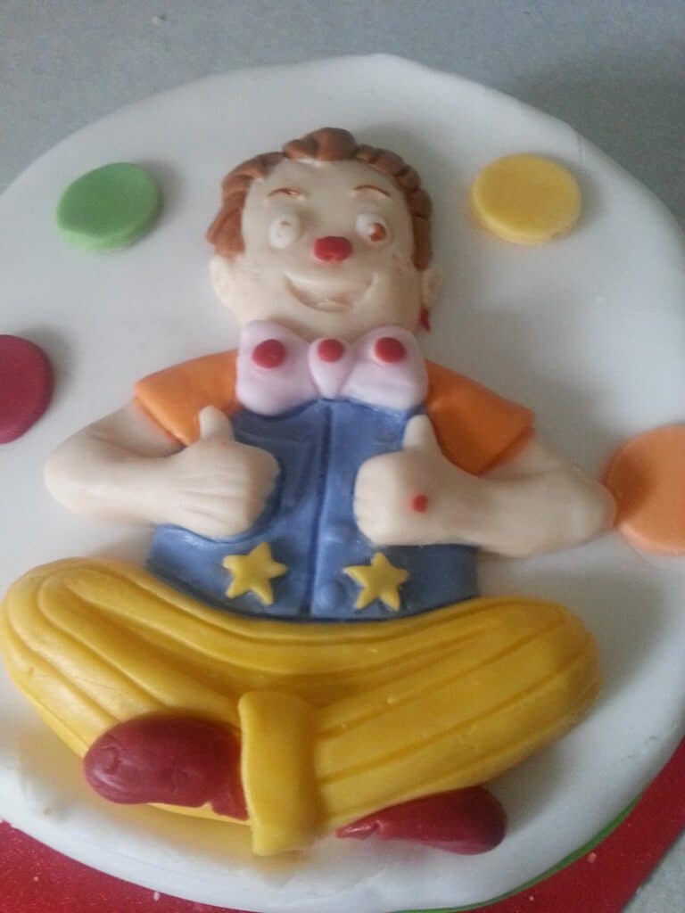 A fondant clown decoration sits cross-legged on a white surface, with yellow trousers, red shoes, a blue waistcoat with yellow stars, orange sleeves, and a red nose. The clown is smiling and giving two thumbs up. Colourful fondant circles decorate the background.
