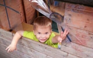 A young child with short light brown hair and wearing a bright green shirt smiles and reaches out playfully through a window of a wooden playhouse. The child’s upper body is visible above the wooden window ledge.