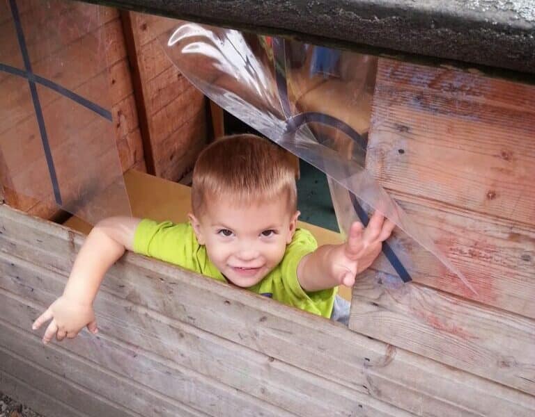 A young child with short light brown hair and wearing a bright green shirt smiles and reaches out playfully through a window of a wooden playhouse. The child’s upper body is visible above the wooden window ledge.