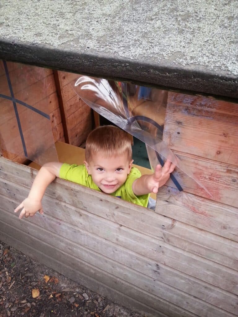 A young child in a bright green shirt smiles and reaches out through a window of a wooden playhouse. The playhouse has a rough, grey roof and clear plastic covering the window. The child is looking up and appears happy and playful.