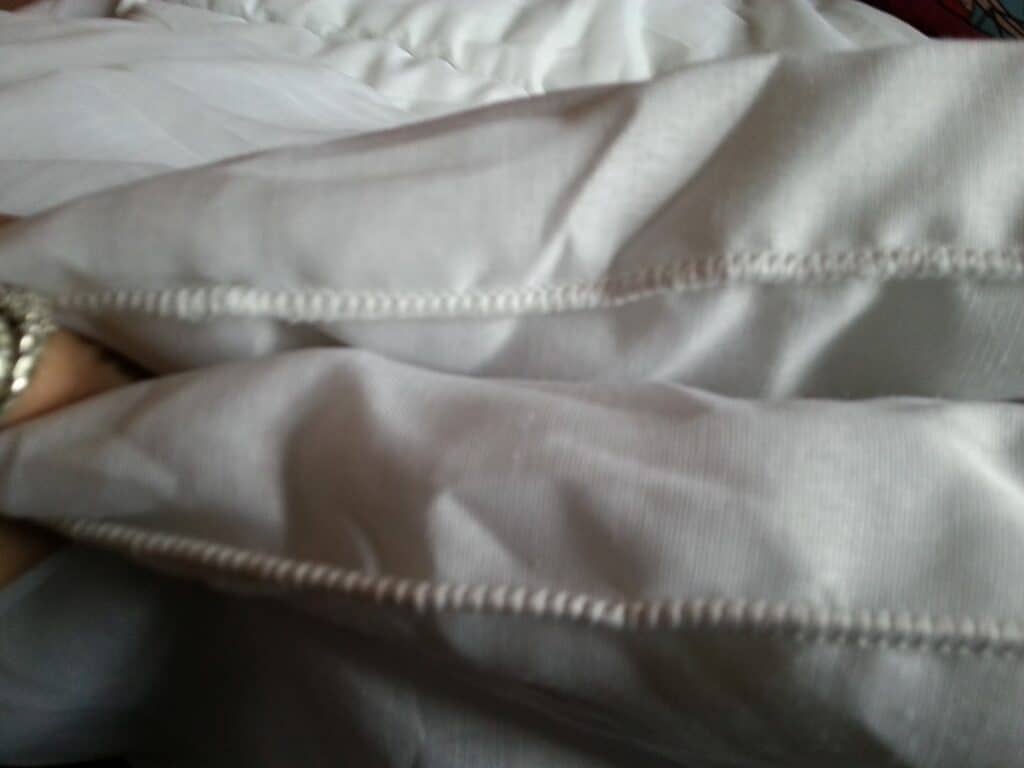 Close-up of two white pillows with visible stitched seams, placed next to each other on a white bed. The stitching forms a line along the edge of the pillows, and a person's hand wearing a bracelet is partially visible holding one pillow.