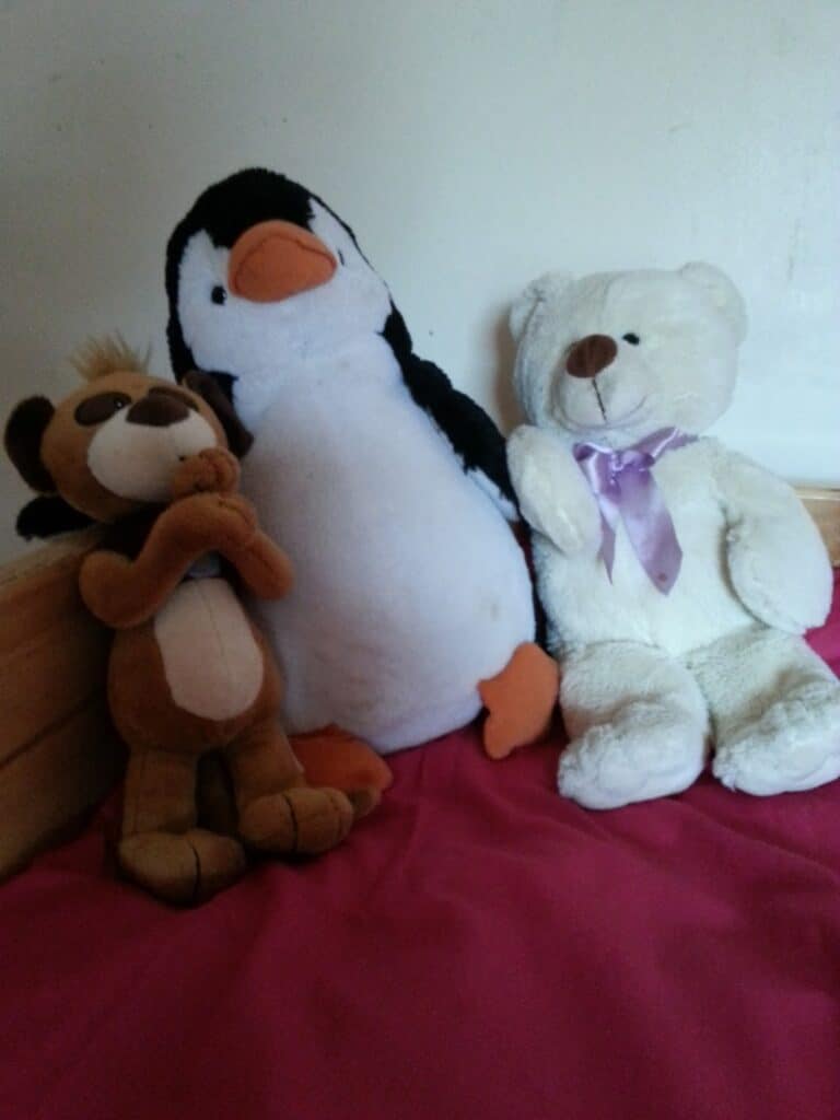 Three soft toys sit on a pink blanket: a brown monkey with hands clasped, a black and white penguin with orange feet and beak, and a white teddy bear with a purple ribbon around its neck, all against a plain light wall.