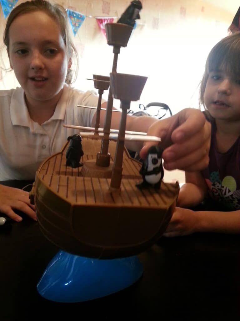 Two children sit at a table playing with a brown plastic pirate ship toy set on a blue base. One child is moving a small pirate figure, whilst the other looks on. Colourful triangle bunting hangs in the background.