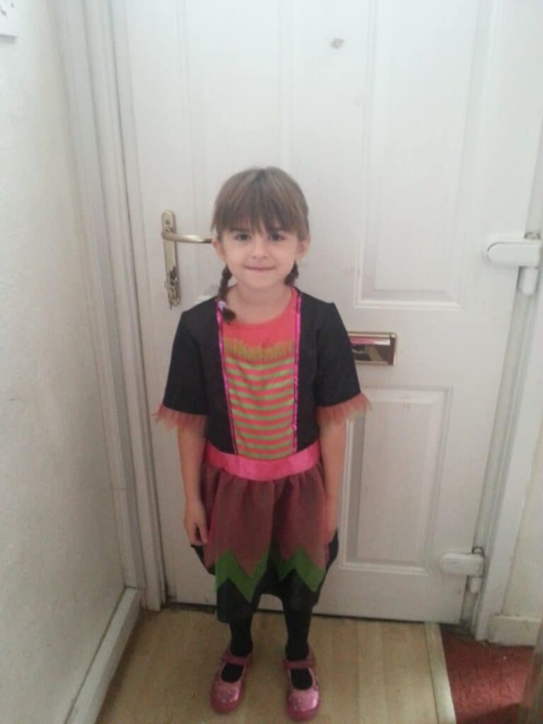 A young girl with brown hair in plaits stands smiling in front of a white door. She wears a colourful costume with a striped bodice, pink collar, black short sleeves, a layered skirt with zigzag hem, black leggings, and shiny pink shoes.