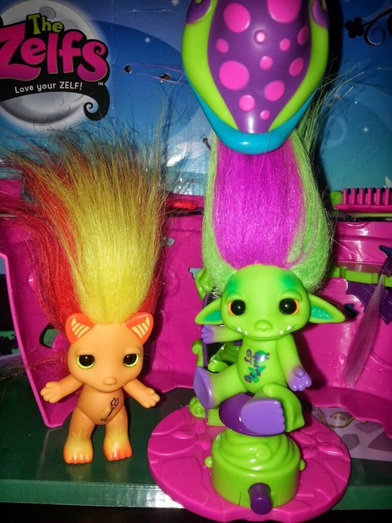 Two colourful toy figures from "The Zelfs" series stand in a vibrant playset. The left figure is orange with a cat-like face and tall yellow hair. The right figure is green with large ears, purple spots, and tall green and purple hair, sitting on a play salon chair.