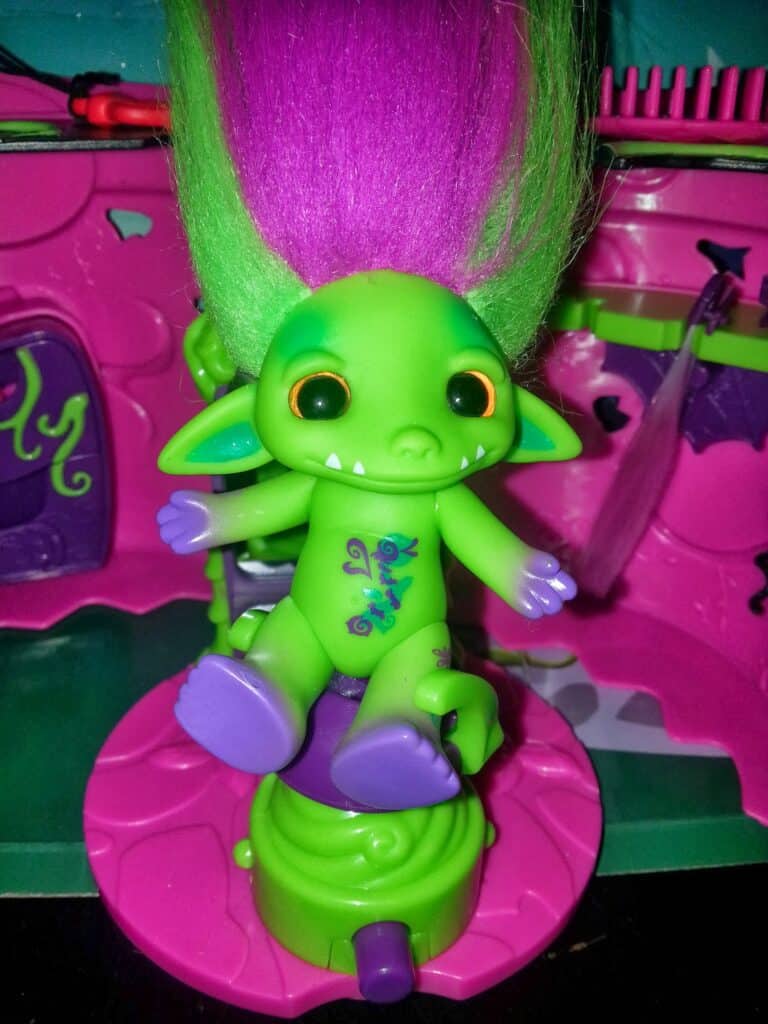 A green troll doll with big orange eyes, pointy ears, a wide smile with small fangs, and long, upright purple and green hair, sits on a pink and green toy chair. The background has pink walls and toy accessories. The troll has purple hands, feet, and a chest design.