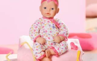 A baby doll wearing a patterned white sleepsuit and a pink headband sits on a folded pink towel. In front, there are pink and yellow toy medical instruments including a stethoscope, spoon, and heart-shaped accessory. The background is pastel pink.