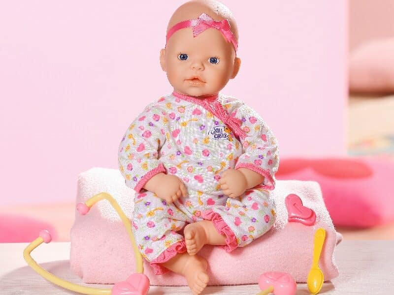 A baby doll wearing a patterned white sleepsuit and a pink headband sits on a folded pink towel. In front, there are pink and yellow toy medical instruments including a stethoscope, spoon, and heart-shaped accessory. The background is pastel pink.