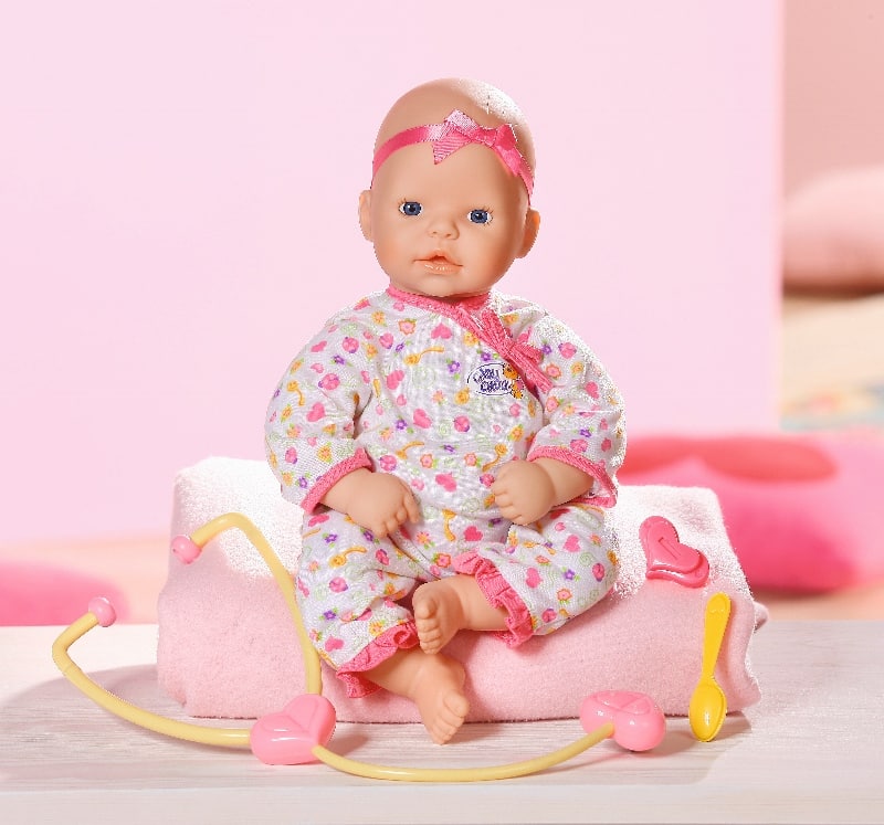 A baby doll wearing a patterned white sleepsuit and a pink headband sits on a folded pink towel. In front, there are pink and yellow toy medical instruments including a stethoscope, spoon, and heart-shaped accessory. The background is pastel pink.