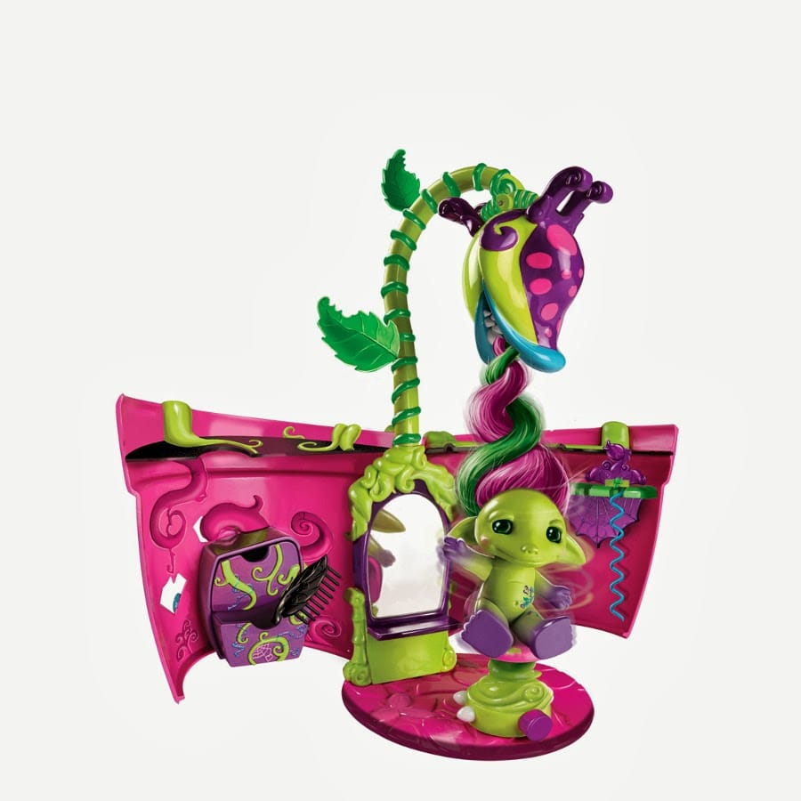 A colourful, whimsical toy salon set features a green creature with purple and pink hair, seated in front of a mirror. The salon has a curved pink backdrop, leafy green plant details, a hanging hairdryer, tiny accessories, and playful purple accents.