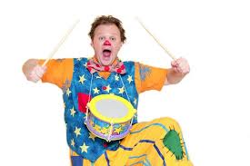 A surprised clown with a red nose, colourful clothes, and a star-patterned waistcoat, holds drumsticks and plays a small toy drum hanging from his neck. His mouth is open wide, and he stands against a plain white background.
