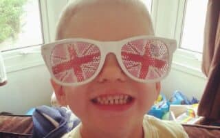 A young boy smiling widely indoors, wearing large white sunglasses with the Union Jack (British flag) design on the lenses. Sunlight streams through a window behind him, and colourful toys are visible in the background.