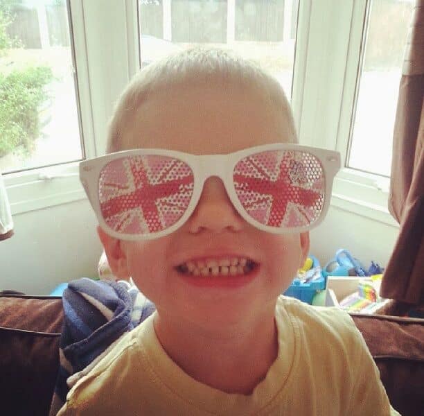A young boy smiling widely indoors, wearing large white sunglasses with the Union Jack (British flag) design on the lenses. Sunlight streams through a window behind him, and colourful toys are visible in the background.