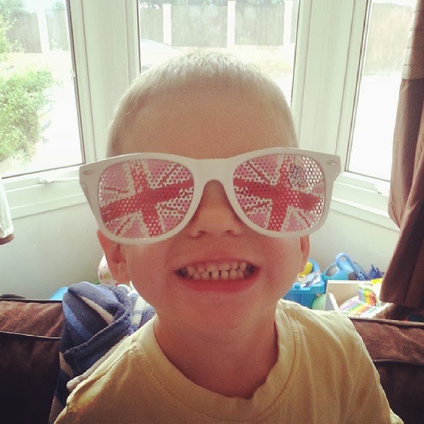 A young boy smiling widely indoors, wearing large white sunglasses with the Union Jack (British flag) design on the lenses. Sunlight streams through a window behind him, and colourful toys are visible in the background.