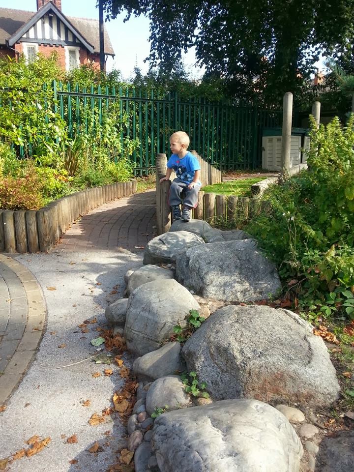 A young boy in a blue shirt and grey trousers sits thoughtfully on a wooden bench in a sunny garden. He is surrounded by green bushes, trees, a stone path, and large rocks. A wooden fence and a house with a gabled roof appear in the background.