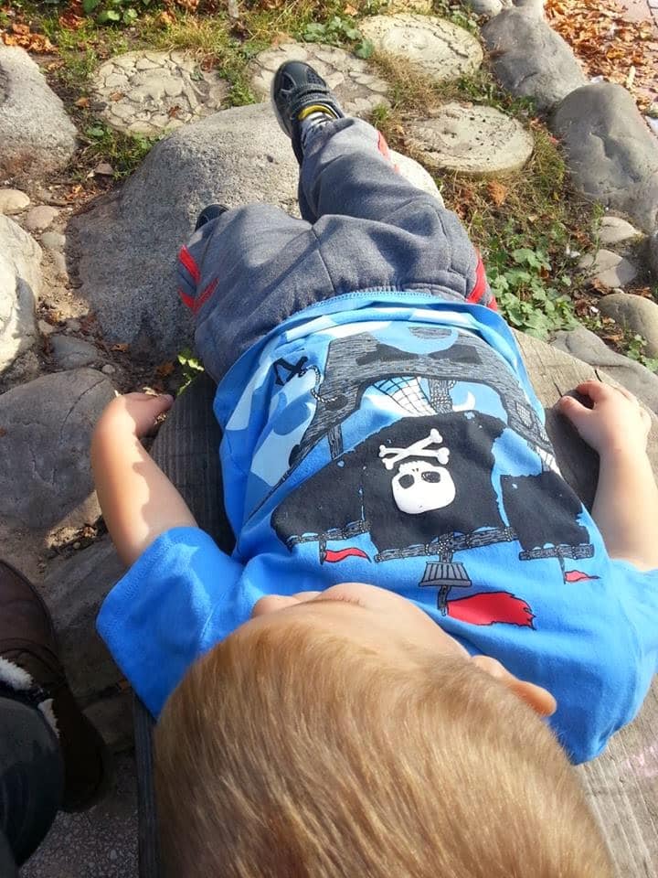 A young child with light brown hair lies on their back on a wooden bench outdoors, wearing a blue T-shirt with a cartoon pirate design and grey trousers. The child looks relaxed, surrounded by rocks, greenery, and sunlight.