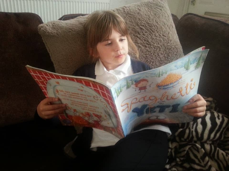 A young child with light brown hair sits on a brown sofa, wearing a white polo shirt and dark cardigan, reading an illustrated children’s book titled “Spaghetti.” The child appears focused, with a zebra-print blanket on their lap.