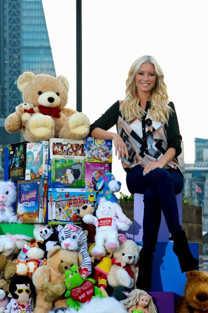 A smiling woman with long blonde hair, dressed in a black blouse and navy trousers, sits on a purple block indoors. She is surrounded by soft toys and colourful board games, with city skyscrapers visible through tall windows behind her.