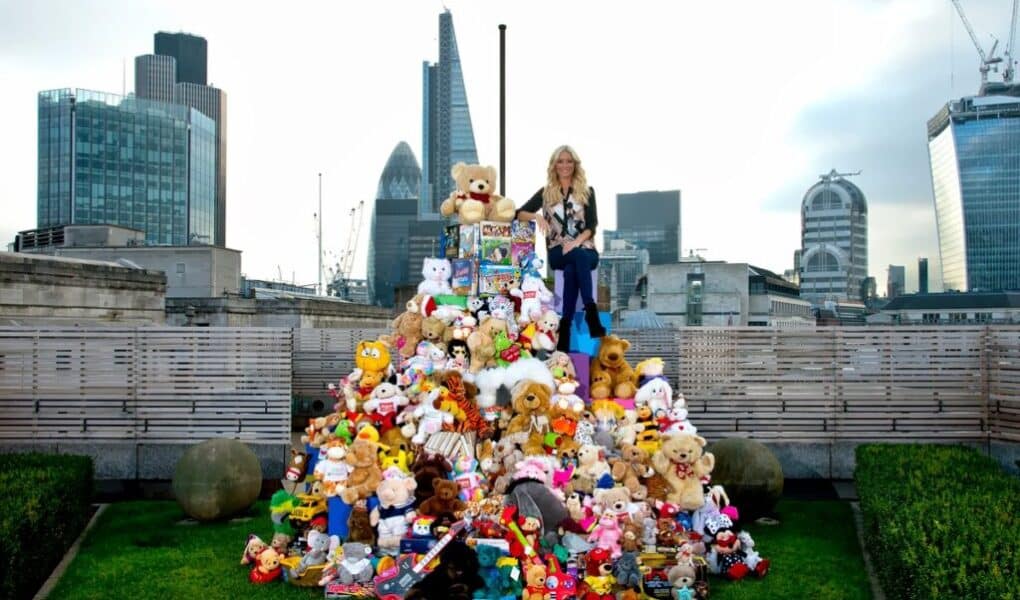 A woman stands on top of a large pyramid of colourful stuffed animals and toys on a rooftop garden, with modern skyscrapers and the Gherkin building visible in the London city skyline behind her. The sky is cloudy and the scene is vibrant and playful.