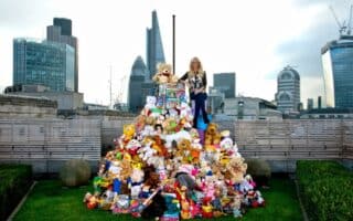 A woman stands on top of a large pyramid of colourful stuffed animals and toys on a rooftop garden, with modern skyscrapers and the Gherkin building visible in the London city skyline behind her. The sky is cloudy and the scene is vibrant and playful.