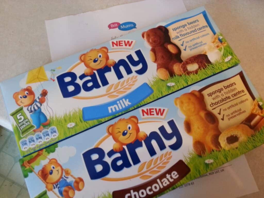 Two boxes of Barny sponge bears are shown, one milk flavour (blue box) and one chocolate flavour (brown box). Each box features a bear mascot, images of the bear-shaped cakes, and text describing the flavoured fillings. A paper is partly visible underneath.