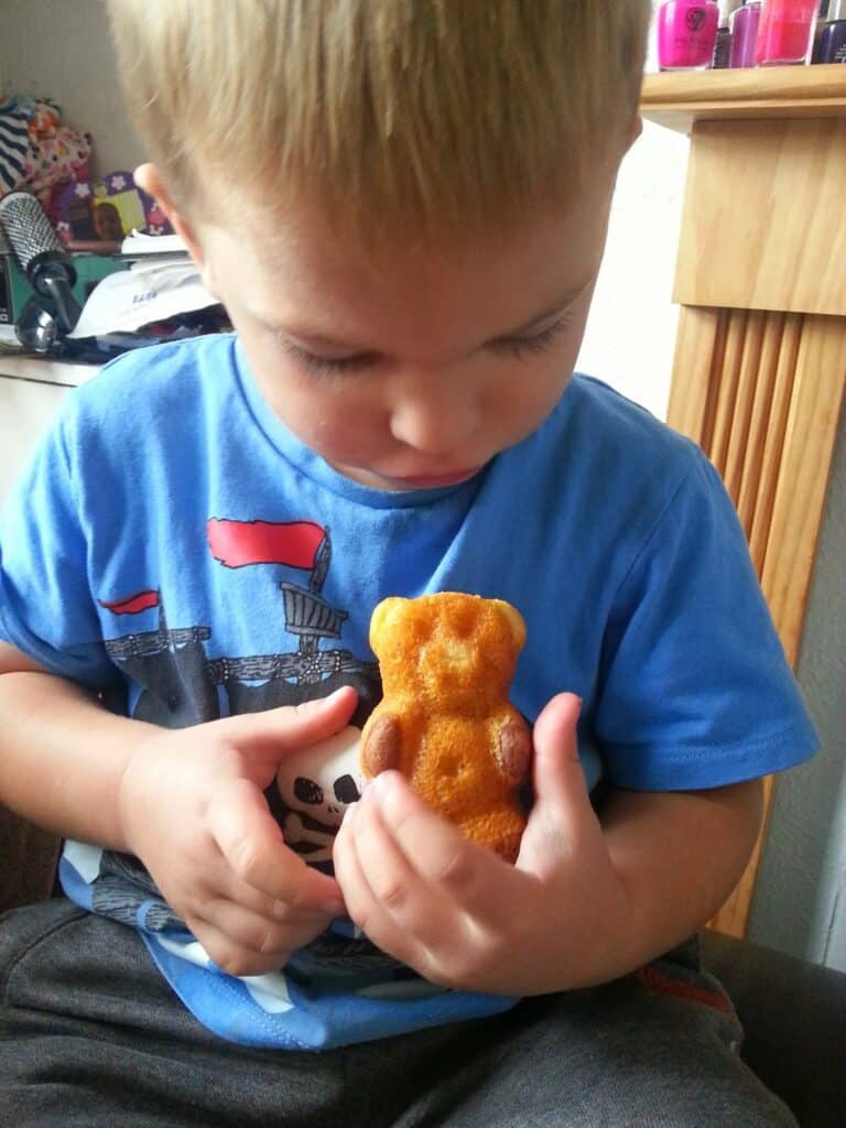 A young child with blonde hair looks down at a bear-shaped cake in their hands. The child wears a blue shirt with a ship graphic and sits indoors, near a fireplace and shelves with colourful items in the background.