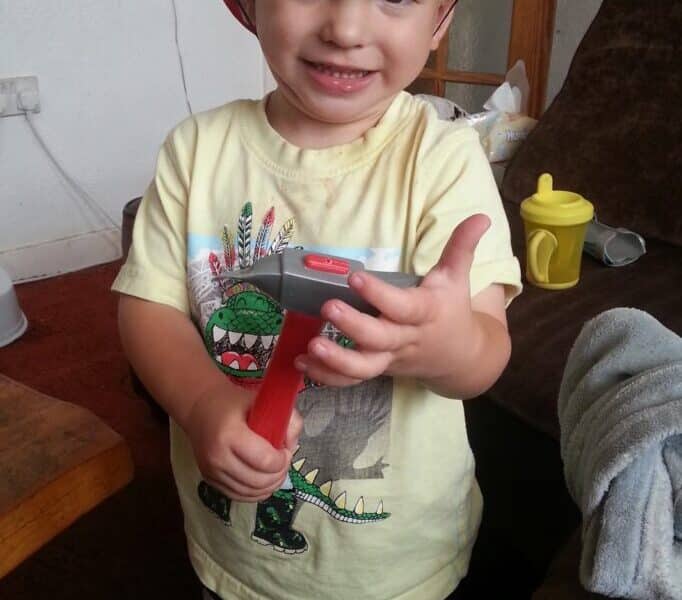 A young child smiles, wearing a red toy fireman’s helmet and holding a toy hammer. The child has light brown hair and is dressed in a yellow T-shirt with a dinosaur graphic. In the background are a brown sofa, blanket, and a yellow beaker.