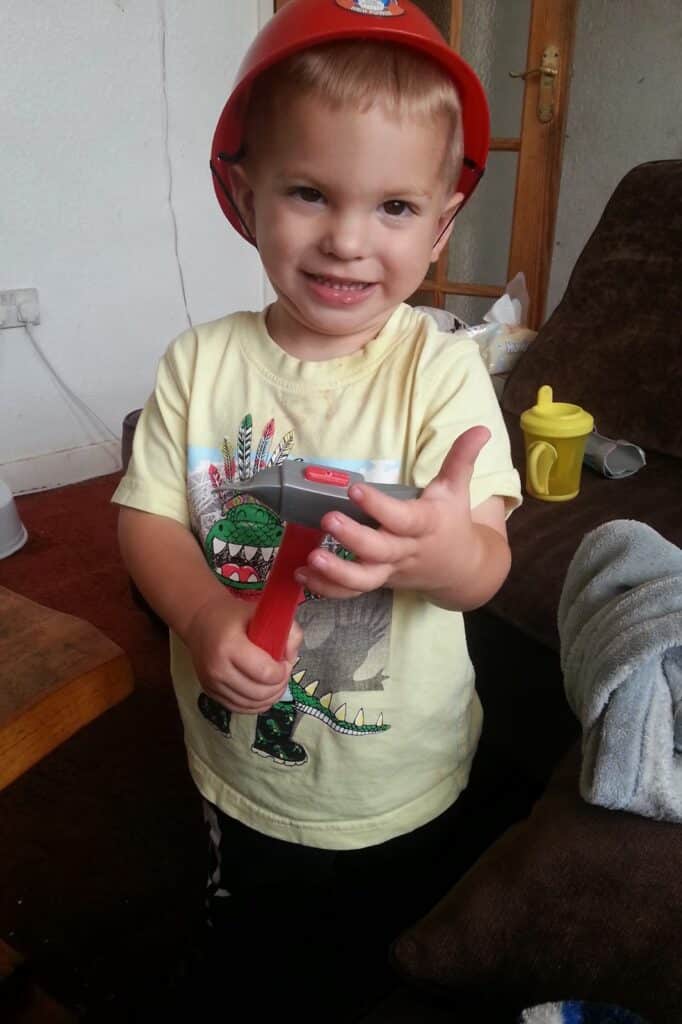 A smiling toddler wearing a red toy fireman’s hat and a yellow shirt with a dinosaur print holds a toy tool. The child stands indoors next to a brown sofa, with a yellow beaker and a blue blanket visible in the background.