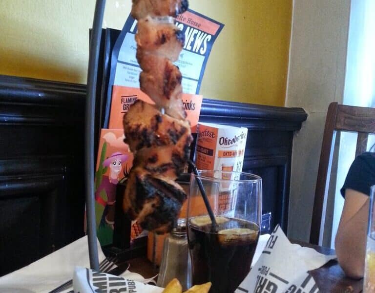 A metal skewer of grilled meat hangs vertically above a basket of golden chips on a restaurant table. Behind, there’s a glass of dark fizzy drink with ice and a straw, menus, and a wood-panelled wall with yellow paint above.