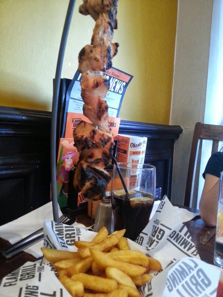 A skewer of grilled meat hangs vertically above a metal tray lined with branded paper, filled with golden chips. A glass of dark fizzy drink with ice and a straw sits beside the food. Menus and a colourful wall are visible in the background.