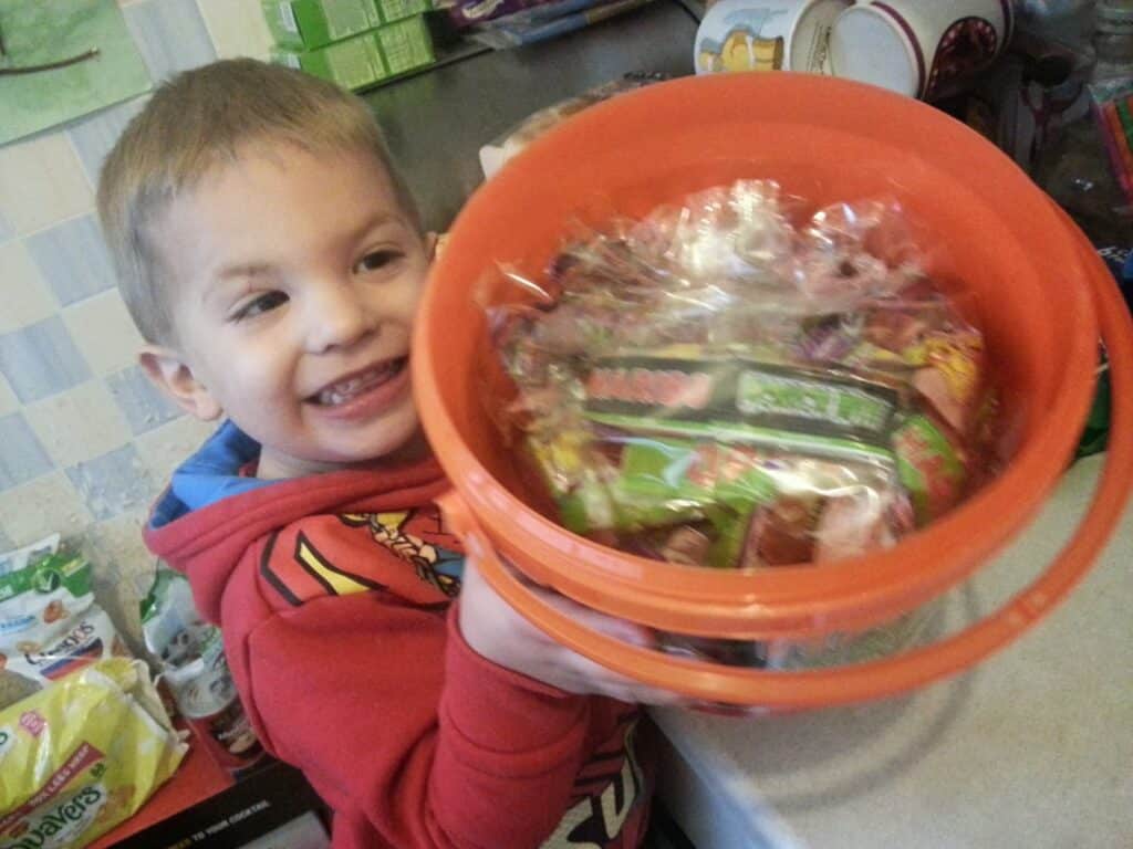 A young child wearing a red jumper smiles whilst holding an orange plastic bucket filled with assorted, colourful wrapped sweets. The background shows a kitchen worktop with bags and household items, suggesting a festive or celebratory occasion.