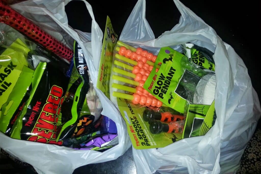 Two white plastic bags filled with Hallowe’en-themed items on a dark surface. Visible are orange glow sticks, a green “Glow Pendant” pack, fake severed fingers, and other assorted spooky sweets and decorations in colourful packaging.