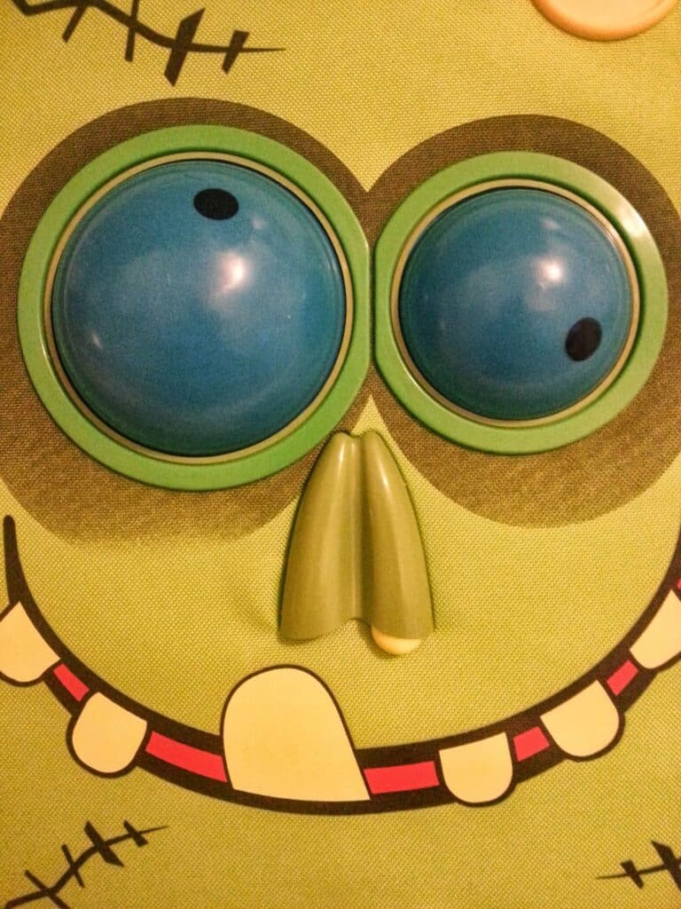 A close-up of a cartoonish green face with two large, blue, bulging eyes, a protruding green nose, a wide smiling mouth with a missing tooth, and black stitched lines resembling scars on the green background.