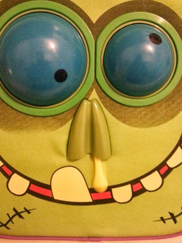 Close-up of a cartoonish green face with large, protruding blue eyes, a wide toothy smile with one big yellow tooth, red gums, a green nose, and a yellow snot hanging from a nostril; the style is playful and exaggerated.