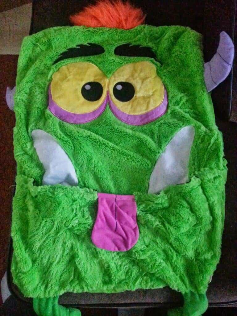 A fuzzy green costume or blanket shaped like a cartoon monster with big yellow and purple eyes, black eyebrows, white felt fangs, a pink tongue, purple horns, and an orange tuft of hair on top, spread out flat on a dark surface.