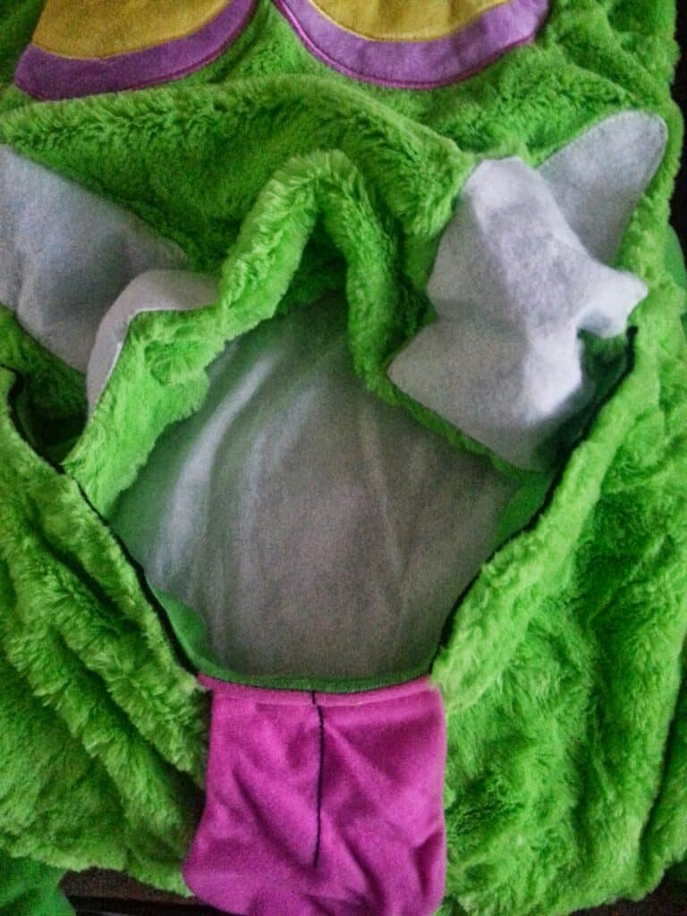 Close-up of a fuzzy green costume with an open mouth lined in white fabric, purple tongue, and white felt teeth. Part of yellow eyes is visible at the top. The texture is soft and plush, resembling a cartoonish or monstrous character.