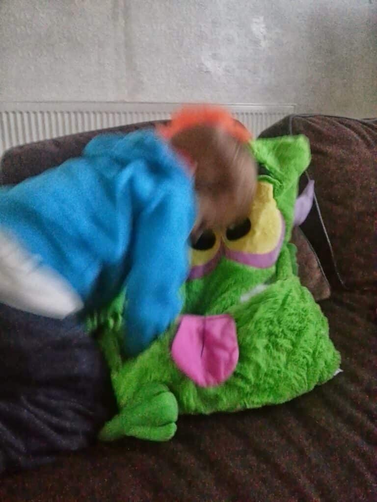 A small child in a bright blue hoodie leans forward on a sofa, appearing to hug or play with a large, green, fuzzy cushion shaped like a cartoon monster with big yellow eyes and a pink tongue. The background shows a radiator and a neutral-coloured wall.