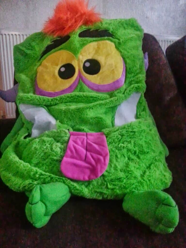 A soft toy shaped like a bright green, cartoonish monster with big yellow eyes, bushy orange eyebrows, a pink tongue sticking out, white teeth, and floppy green arms and feet. It sits on a dark surface with a radiator in the background.
