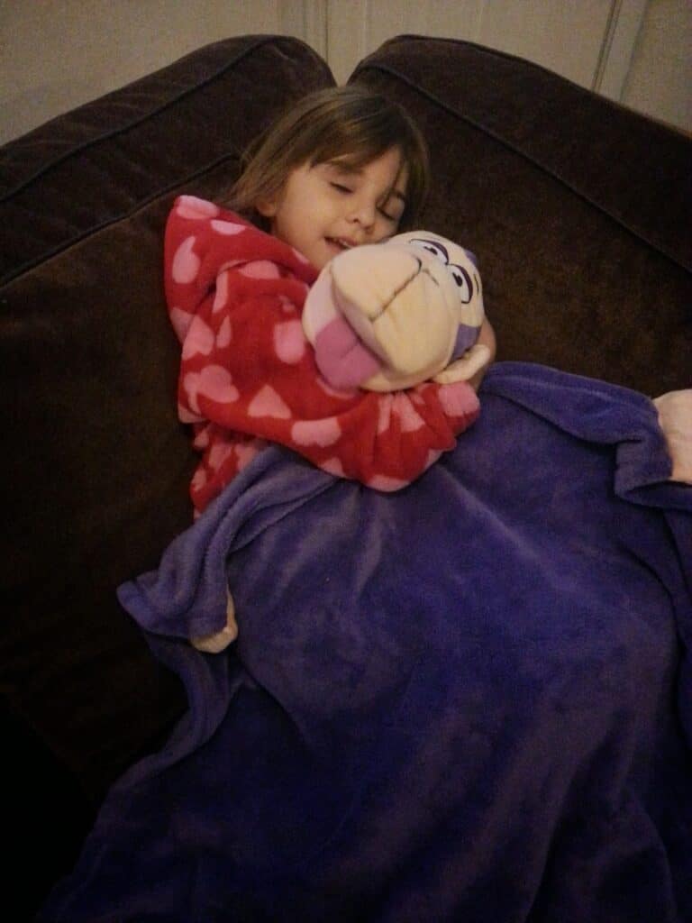 A young child in a red dressing gown with pink hearts lies on a brown sofa, hugging a cartoon monkey soft toy. She is covered with a purple blanket and appears to be resting or sleeping, with eyes mostly closed and a peaceful expression.