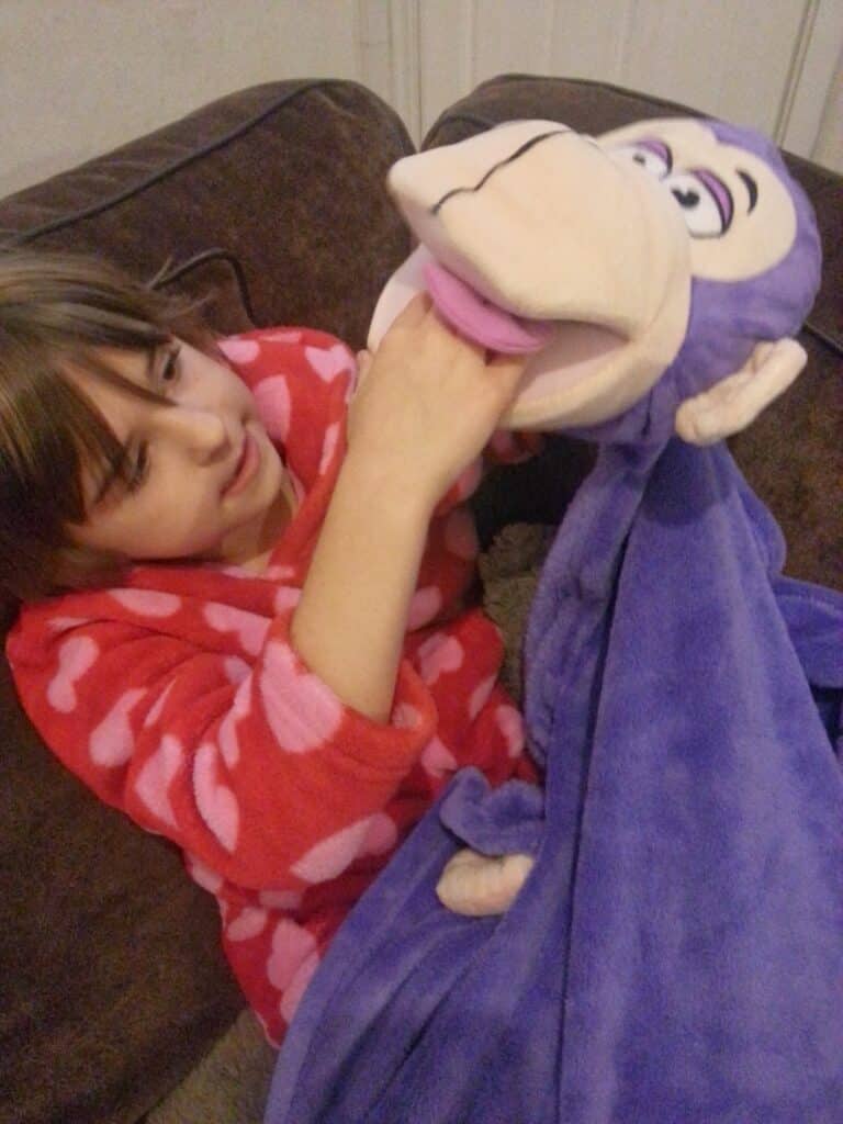 A young girl in a red dressing gown with pink hearts sits on a brown sofa, playing with a large purple monkey puppet. The girl’s hand is inside the puppet’s mouth, making it open, and she looks at the puppet with interest.