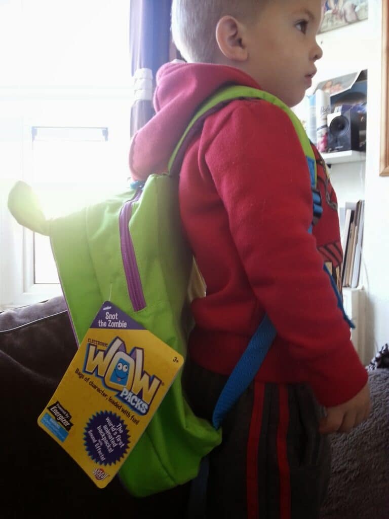 A young child in a red hoodie and striped trousers stands sideways indoors, wearing a green rucksack. Attached to the rucksack is a yellow tag labelled "WOW Packs." The child looks forwards, and sunlight comes through a window in the background.