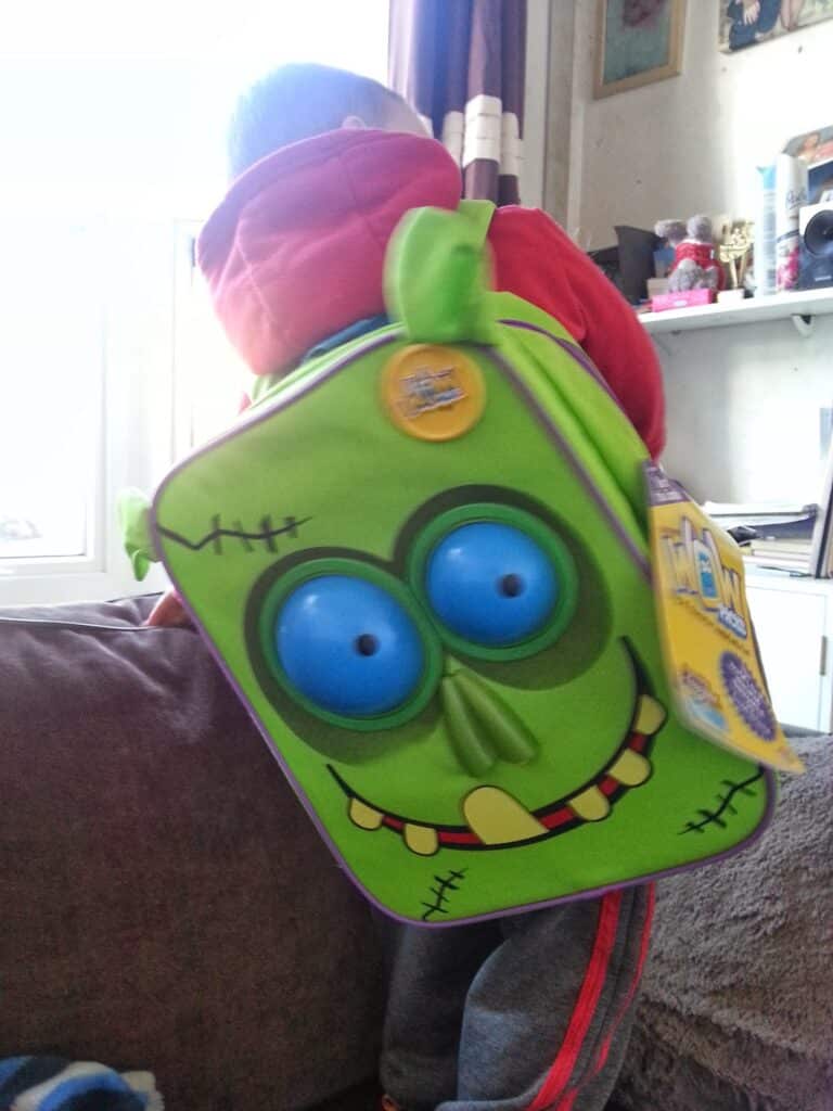 A young child in a red hoodie, facing away, wears a bright green, monster-themed rucksack with a big blue-eyed cartoon face and a toothy grin. The child is standing on a sofa in a sunlit room with shelves and framed pictures in the background.