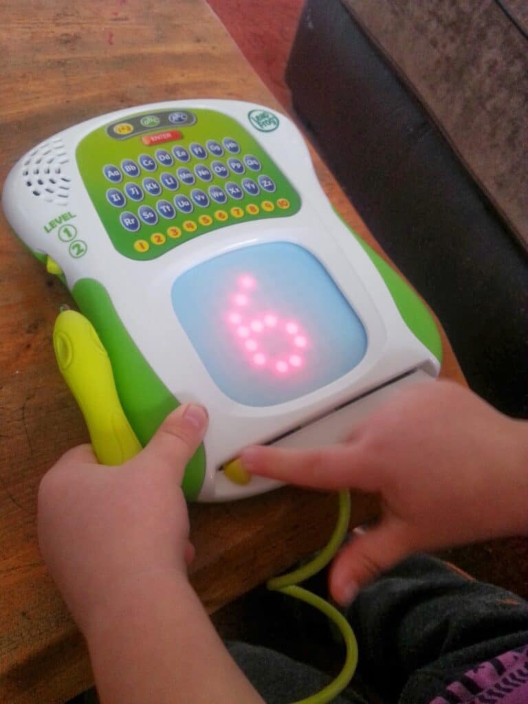 A child uses a LeapFrog educational tablet, pressing a yellow button with one hand. The screen displays a large, glowing number 6 in red dots. The device has a colourful alphabet keypad and sits on a wooden surface.