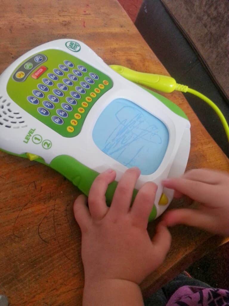 A child’s hands operate a LeapFrog educational toy tablet on a wooden surface. The screen displays a line drawing of an aeroplane, and the child is pressing a yellow button while holding the toy’s green stylus, which is attached by a cord.