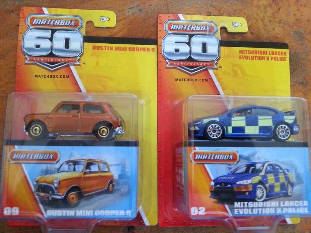 Two Matchbox 60th Anniversary toy cars in packaging: a brown Austin Mini Cooper S (left) and a blue-and-yellow chequered Mitsubishi Lancer Evolution X Police car (right), both on a brown surface. Packaging features car images and branding.