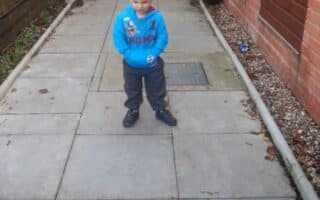 A young boy with short hair stands on a paved walkway outdoors, wearing a blue hoodie and grey trousers. He is looking down and smiling with his hands clasped in front. The path is bordered by a brick building, plants, and a low wall.