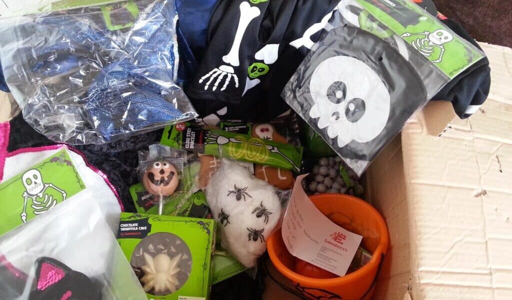 A cardboard box filled with Halloween-themed items, including skeleton costumes, a pumpkin bucket, fake spider webs with plastic spiders, chocolate treats, and a black bag with a white skull design. Packaging and a shopping receipt are also visible.